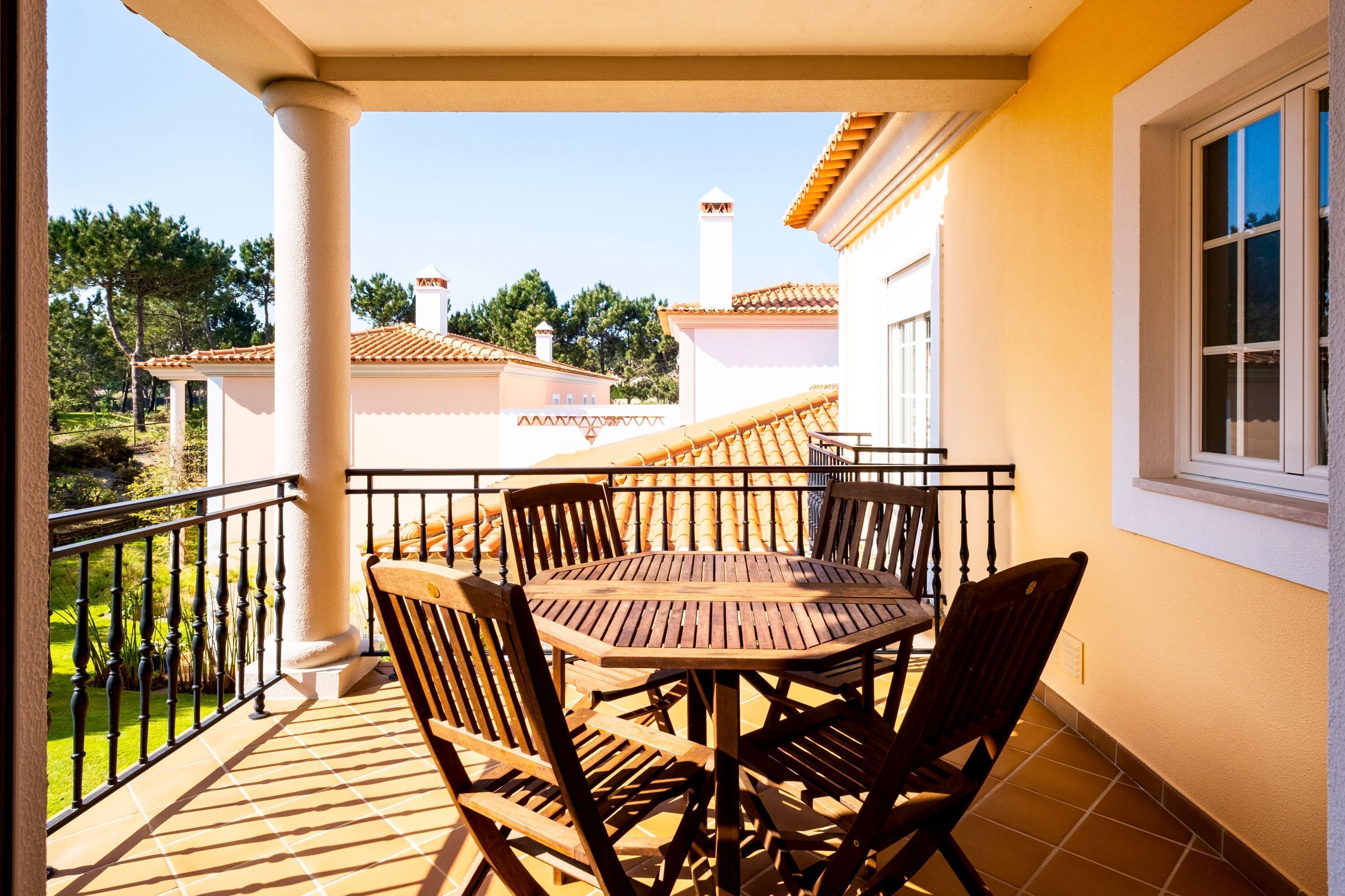 Un balcón con mesa y sillas de madera, piso de baldosas y vista a casas vecinas.