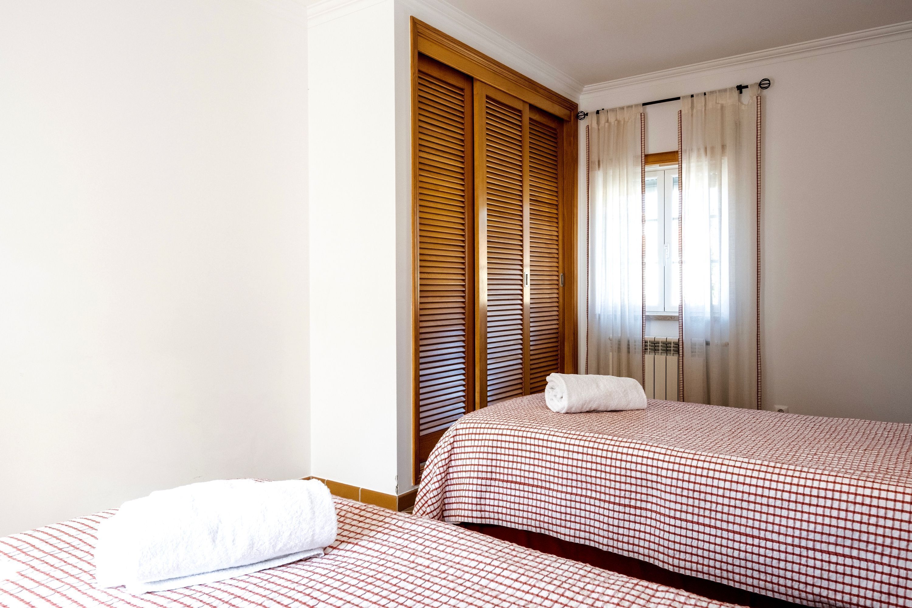 Habitación de hotel con dos camas, contraventanas de madera, un ventanal con cortinas y una toalla blanca sobre una de las camas.