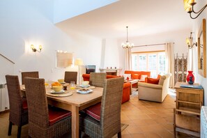 A dining area with a wooden table and wicker chairs, a living room with a sofa and a TV, and a kitchen in the background.