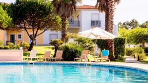 A swimming pool with lounge chairs and umbrellas, surrounded by palm trees and a building with balconies.