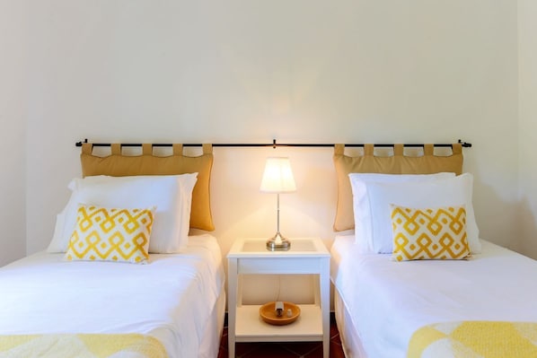 A hotel room with two beds, a bedside table with a lamp, and a small shelf.