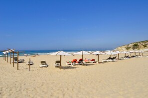 Ein Strand mit Reihen von Liegestühlen und Sonnenschirmen, einem klaren blauen Himmel und dem Meer im Hintergrund.
