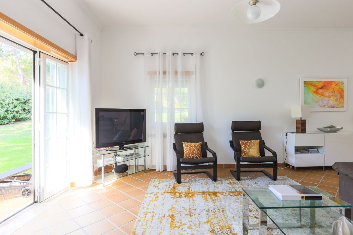 A living room with a TV, two armchairs, a glass coffee table, and a view of a garden through a sliding door.