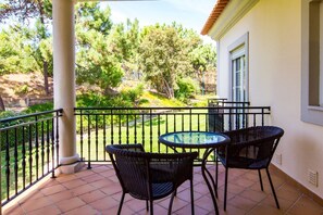 A balcony with a glass table and two wicker chairs, overlooking a garden with trees and a lawn.
