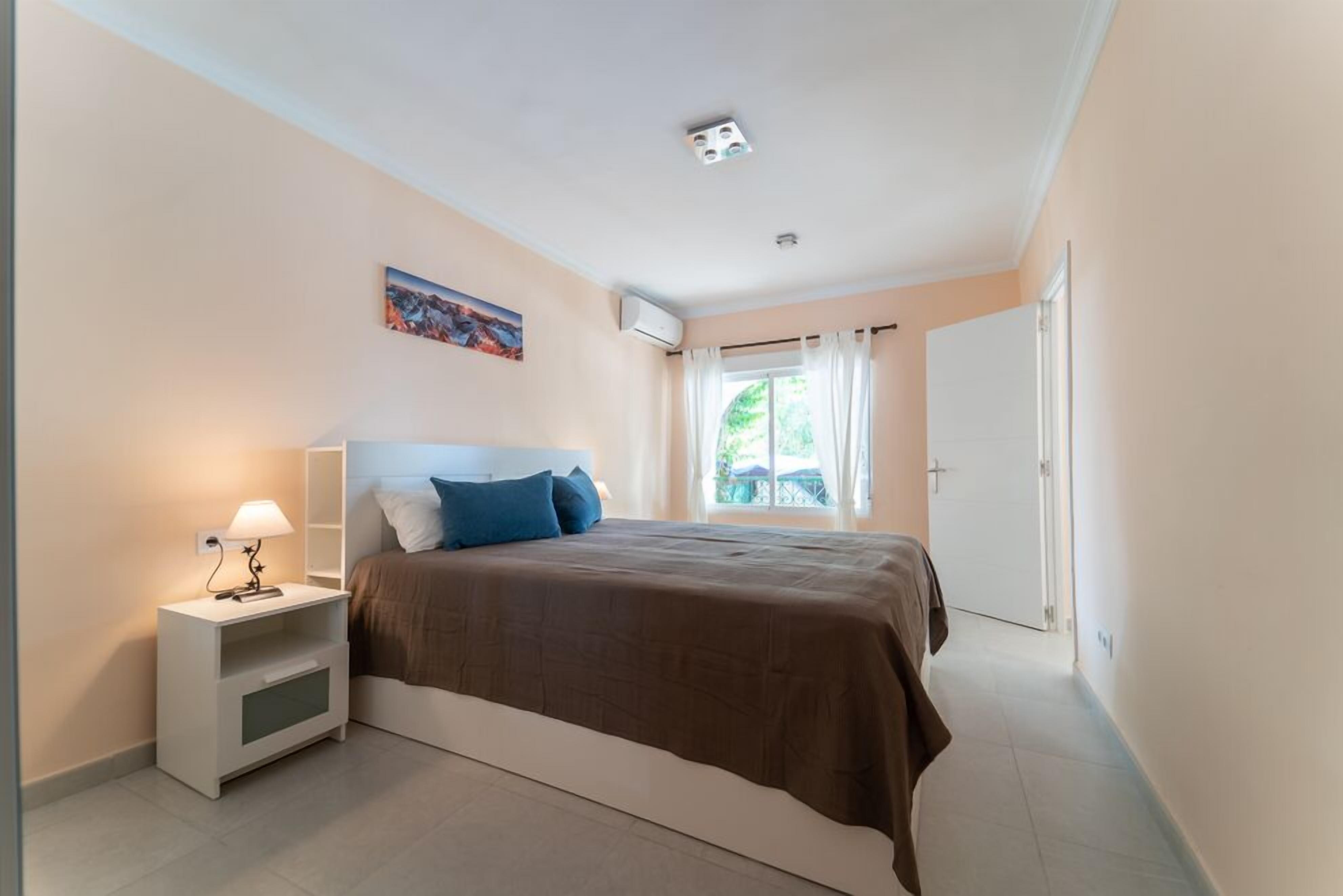 Une chambre à coucher comprenant un lit, une table de chevet, une lampe, un appareil de climatisation et une fenêtre avec des rideaux.