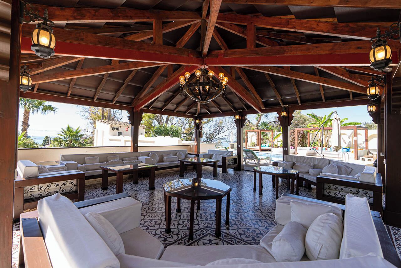 A covered outdoor seating area with white sofas and tables, a patterned floor, and a wooden ceiling structure.