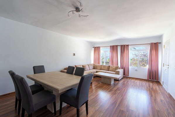 Amplia sala de estar con una mesa de comedor de madera, sillas tapizadas en gris, un sofá color beige y un ventanal con cortinas.