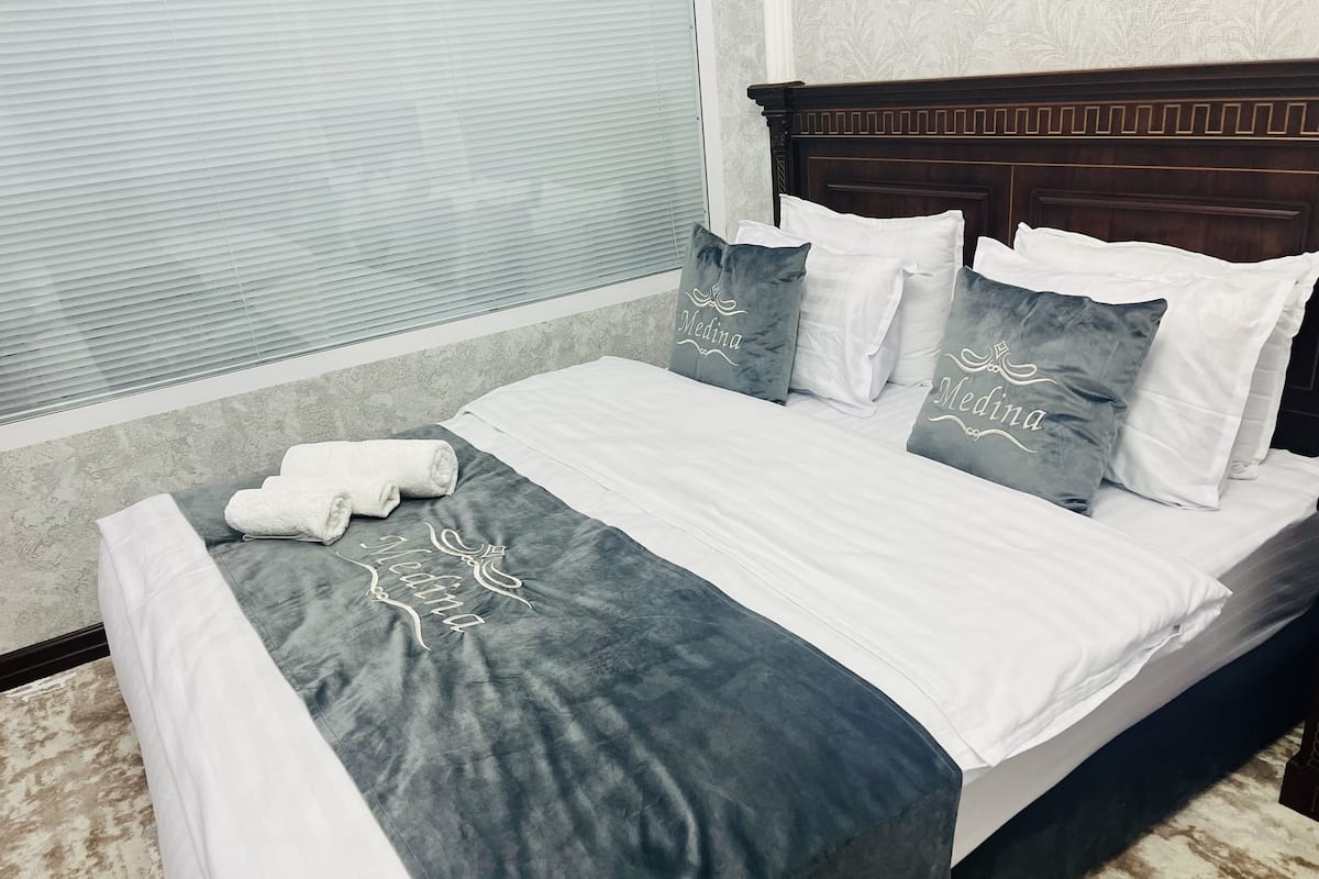 A neatly made bed with a grey and white bedspread, two pillows with the name "Medina," and a rolled white towel on the side.