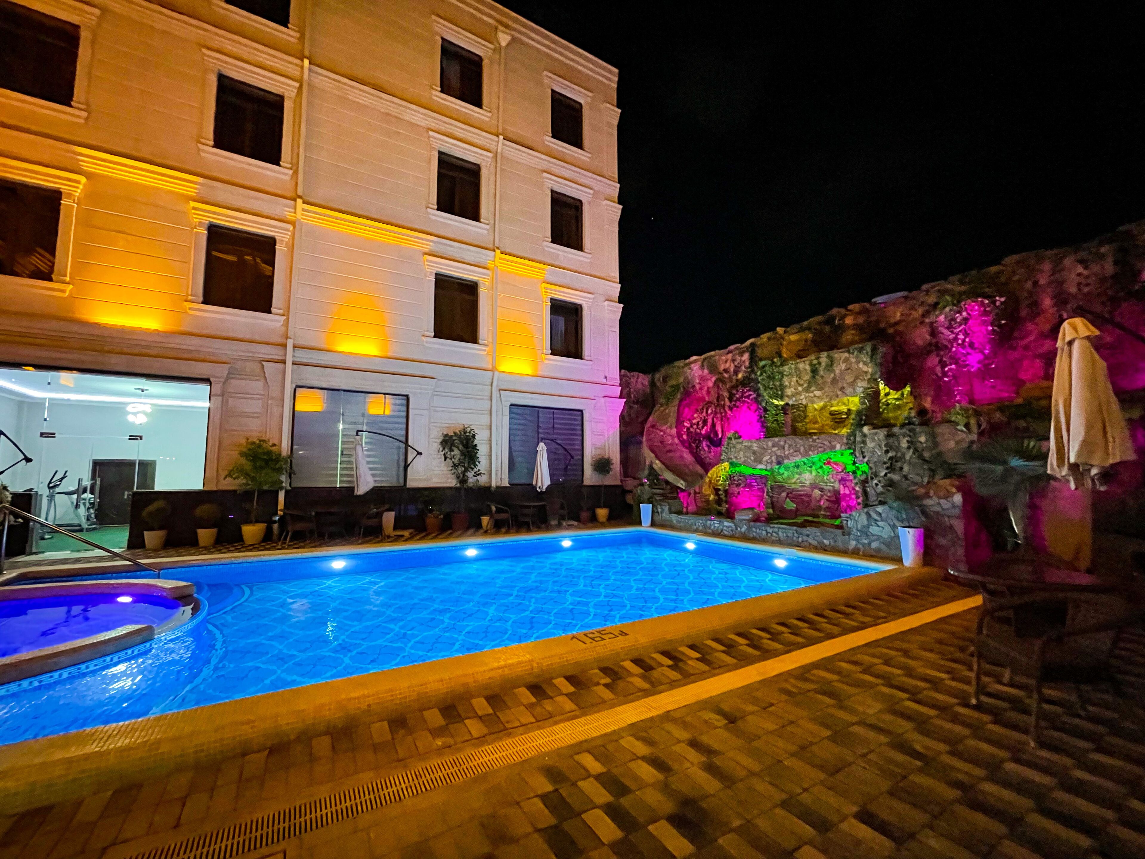 A swimming pool with a tiled deck, a stone wall with colorful lighting, and a building with multiple windows and balconies.