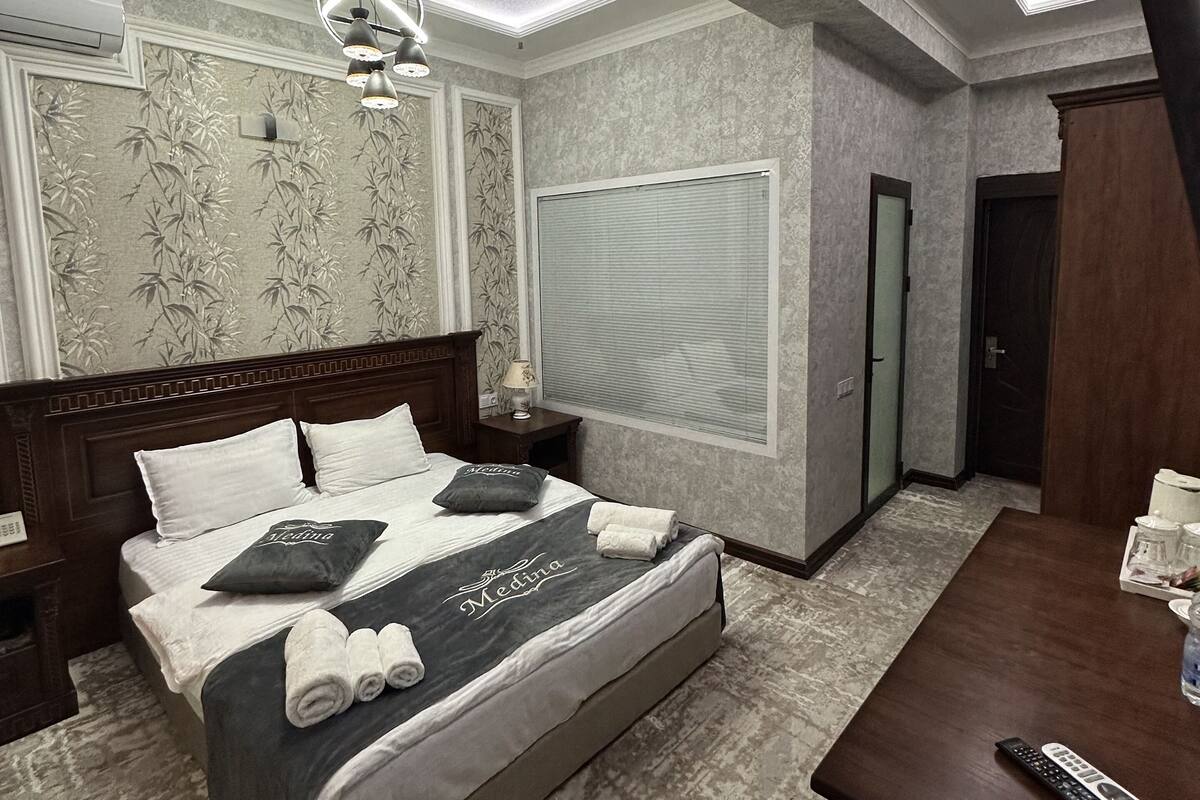 A modern hotel room with a large bed, a wooden nightstand, a desk with a remote control, and a ceiling light fixture.