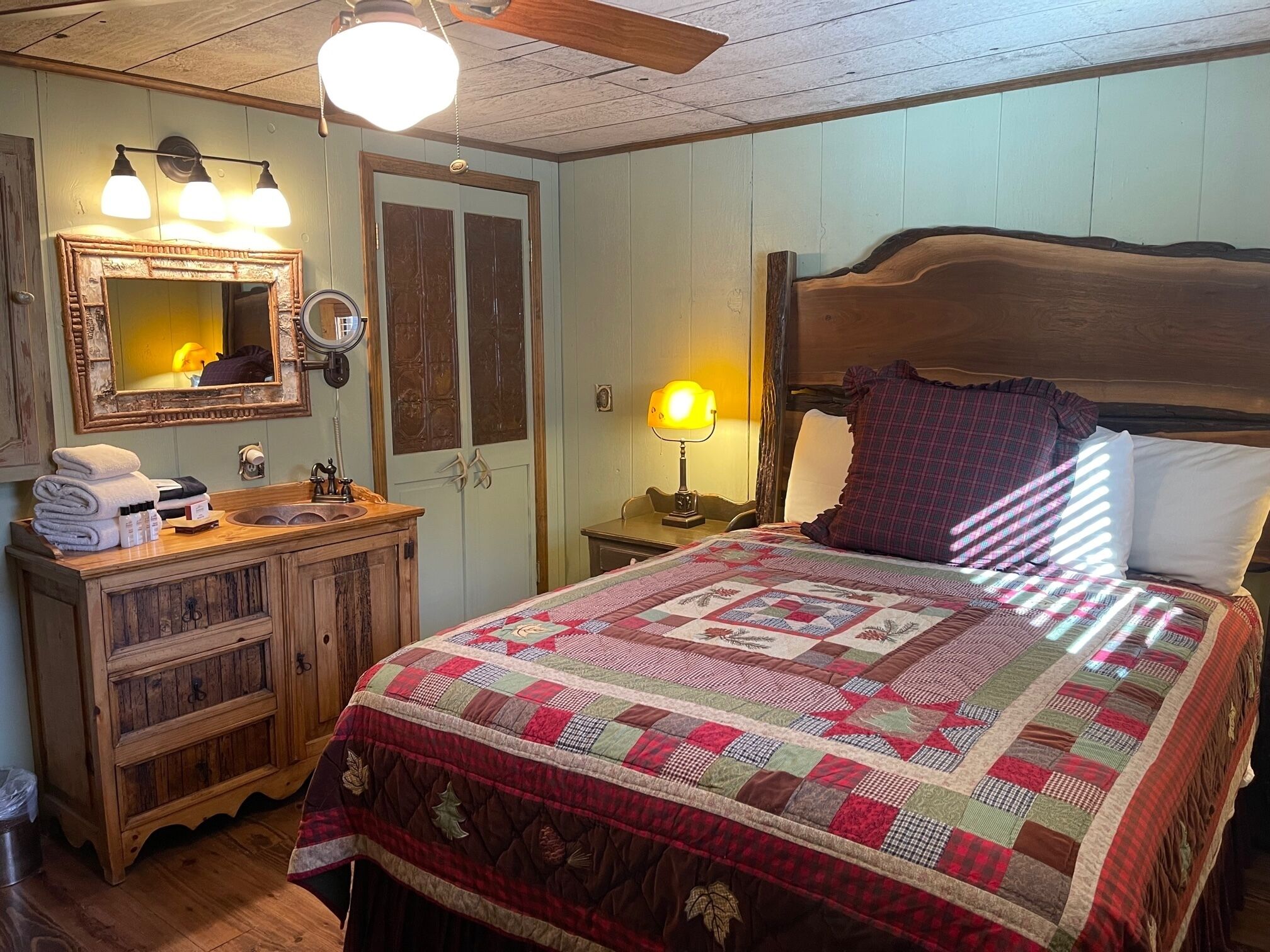 A cozy bedroom with a wooden headboard, a quilted bedspread, a wooden dresser, a mirror, and a lamp.