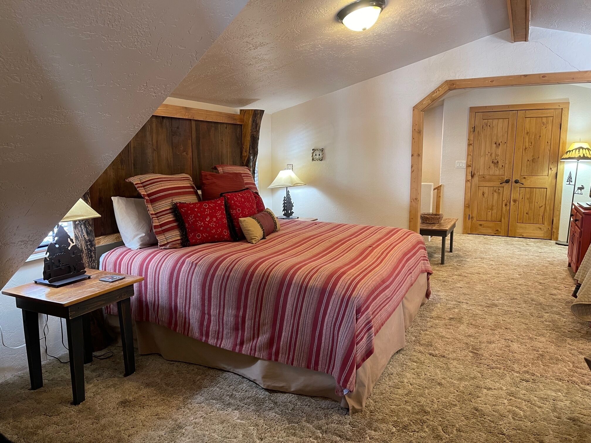 A bedroom with a sloped ceiling, a bed with red and white striped bedding, a nightstand with a lamp, and a wooden wardrobe.
