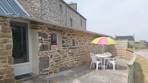 A stone building with a door, a window, and a patio area with a table and chairs. - Small cottage in the countryside, near the sea (Lampaul-Ploudalmézeau)