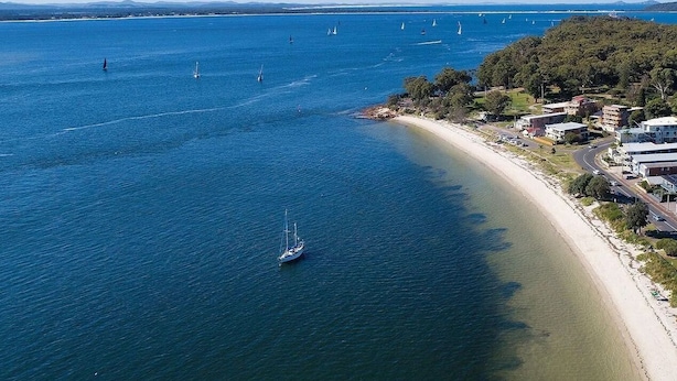An aerial view of a coastal area with a sandy beach, a few sailboats, and buildings along the shore. - Studio 21 - walk to beach marina cafes (Nelson Bay)