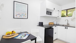 A modern kitchen with white cabinets, a black refrigerator, a microwave, and a sink. There's a framed picture on the wall and a table with fruits and a magazine. - Studio 21 - walk to beach marina cafes (Nelson Bay)