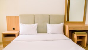 Een net opgemaakt bed met witte lakens, een houten nachtkastje met een lade, een houten bankje met een groene kussen, en een spiegel aan de muur.