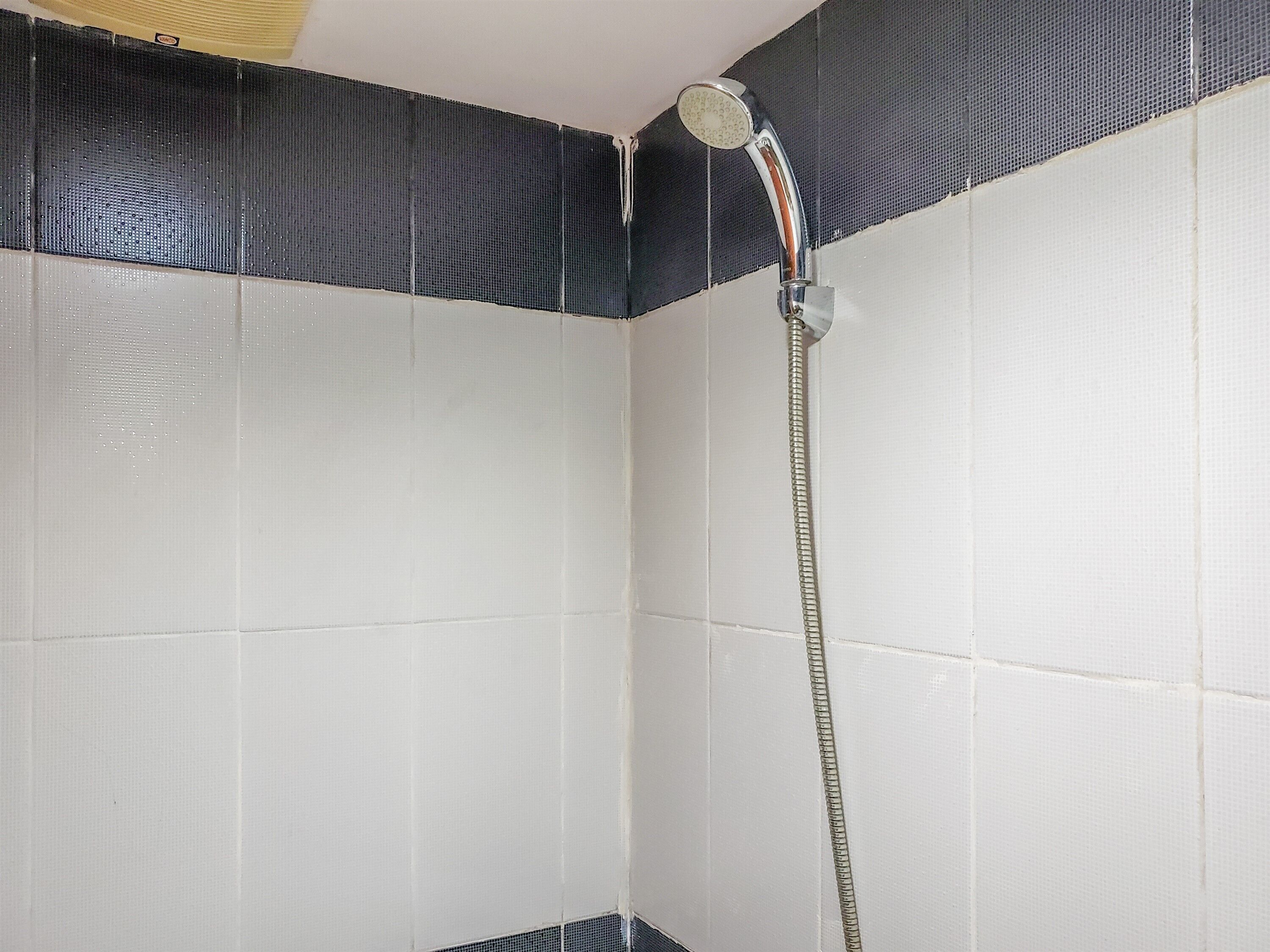 A shower with a handheld hose in a corner of a room with white tiled walls and a black textured ceiling.