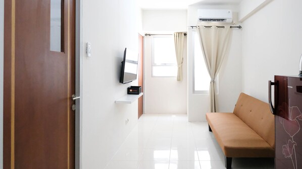 Une pièce moderne et bien éclairée, avec un canapé beige, un téléviseur à écran plat fixé au mur, une petite table sur laquelle se trouve une boîte, et une fenêtre avec des rideaux.