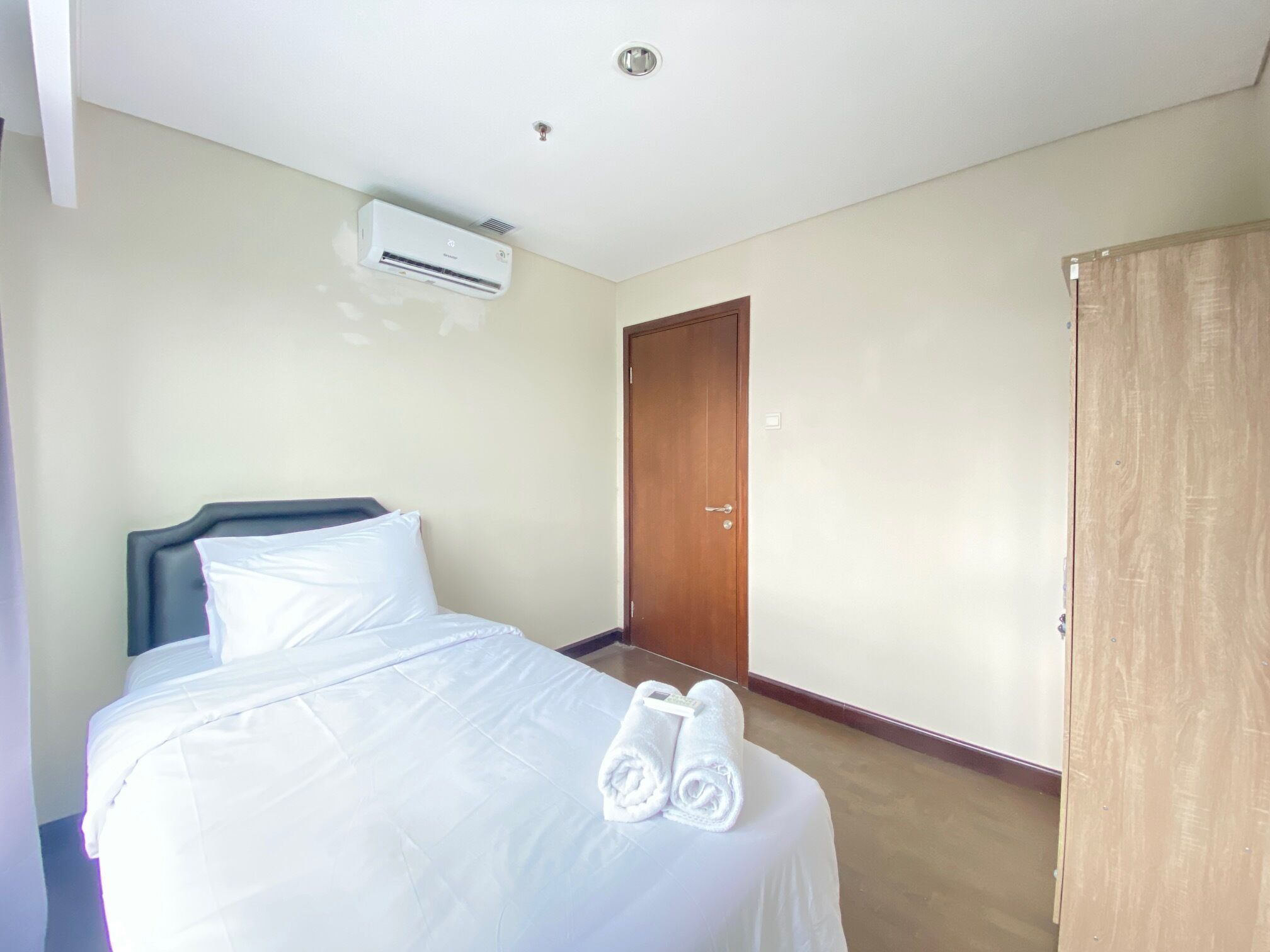 Una camera d'albergo con un letto, un condizionatore, una porta in legno e un asciugamano sul letto.