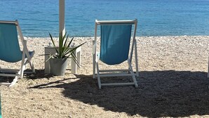 En strand med to stole og et bord under en stråparasol, med udsigt over et roligt hav og bjerge.