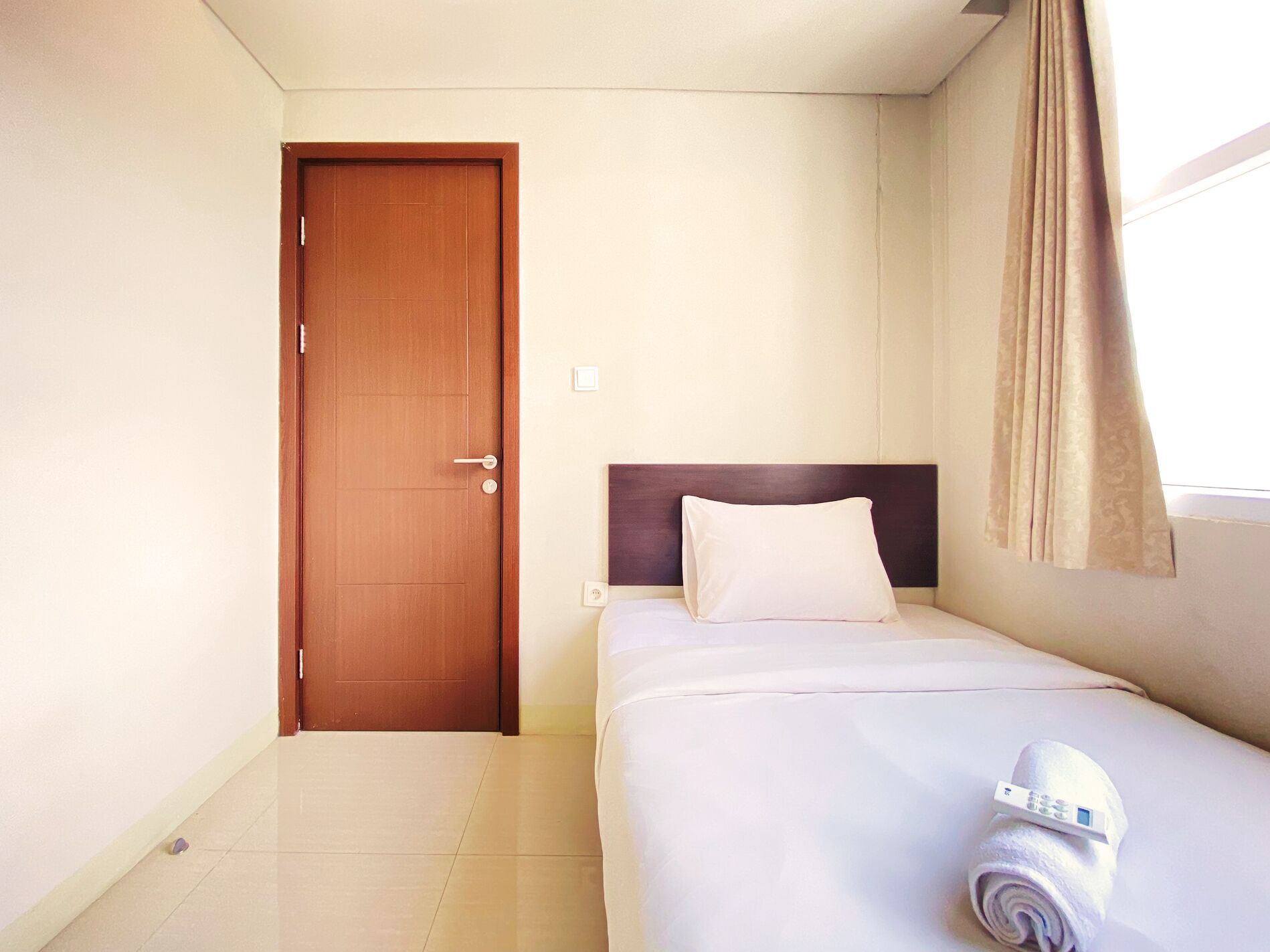 A hotel room with a wooden door, a bed with white bedding, a headboard, and a window with curtains.