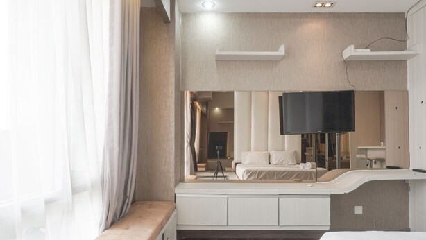 A modern hotel room with a large bed, a built-in bench, a flat-screen TV, and a large window with sheer curtains. - Minimalist And Cozy Studio At Ambassade Kuningan Apartment (Jakarta)