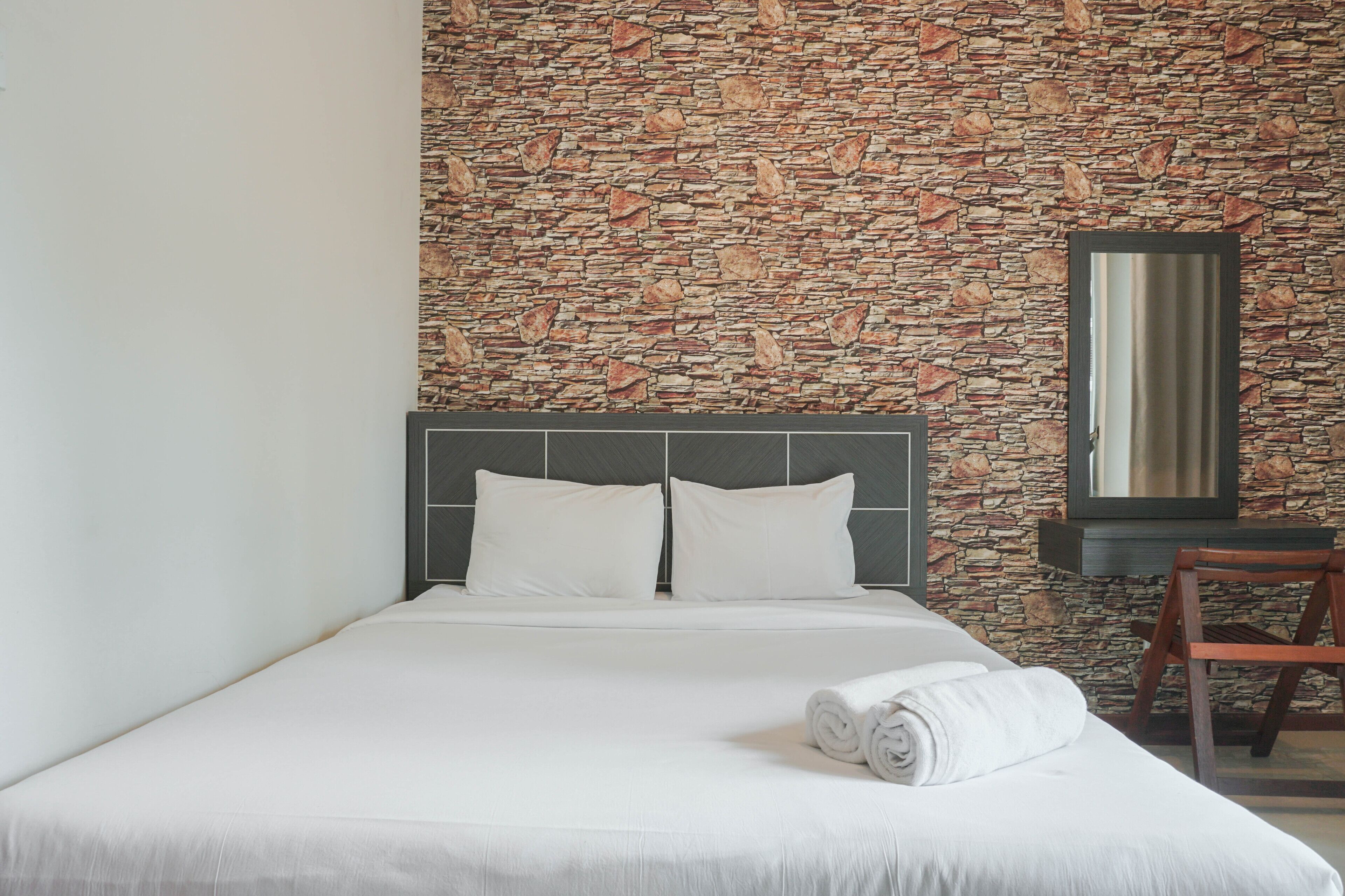 A neatly made bed with white linens and pillows, a stone-patterned wall, and a wooden chair.
