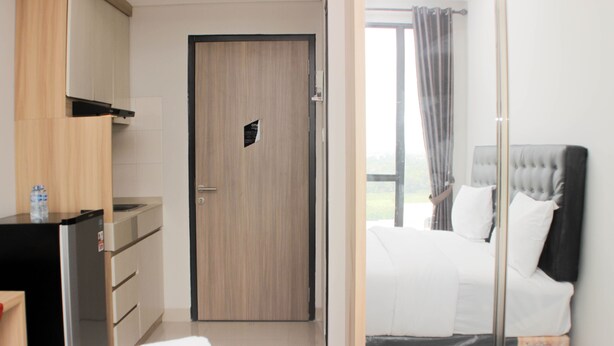 A hotel room with a wooden door, a bed with white bedding, a bedside table, and a view of the outdoors. - Cozy Studio At Monroe Tower Apartment (Cikarang)