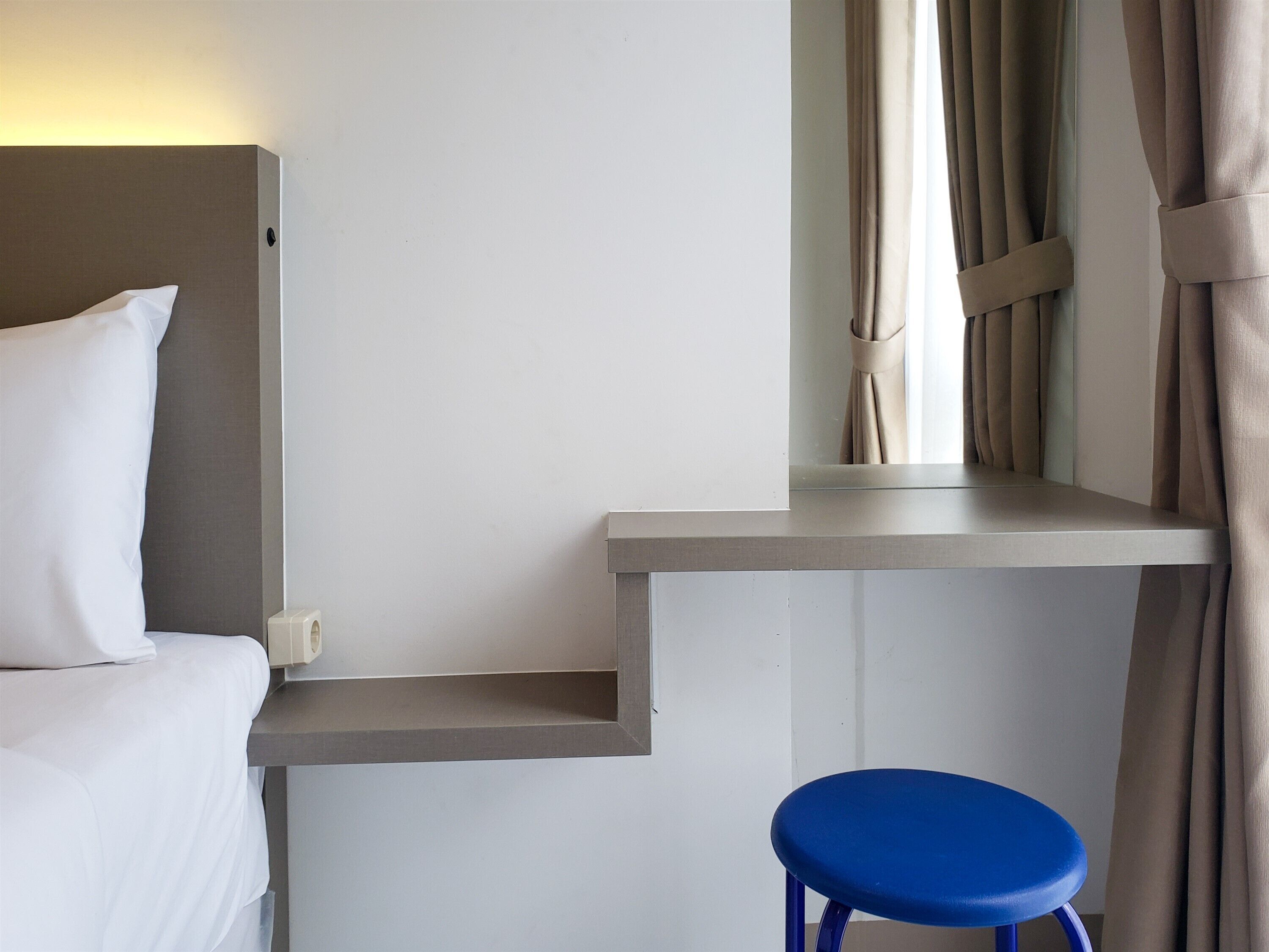 Ein Hotelzimmer mit einem Bett, einem kleinen Schreibtisch, einem blauen Hocker und einem Fenster mit Vorhängen.