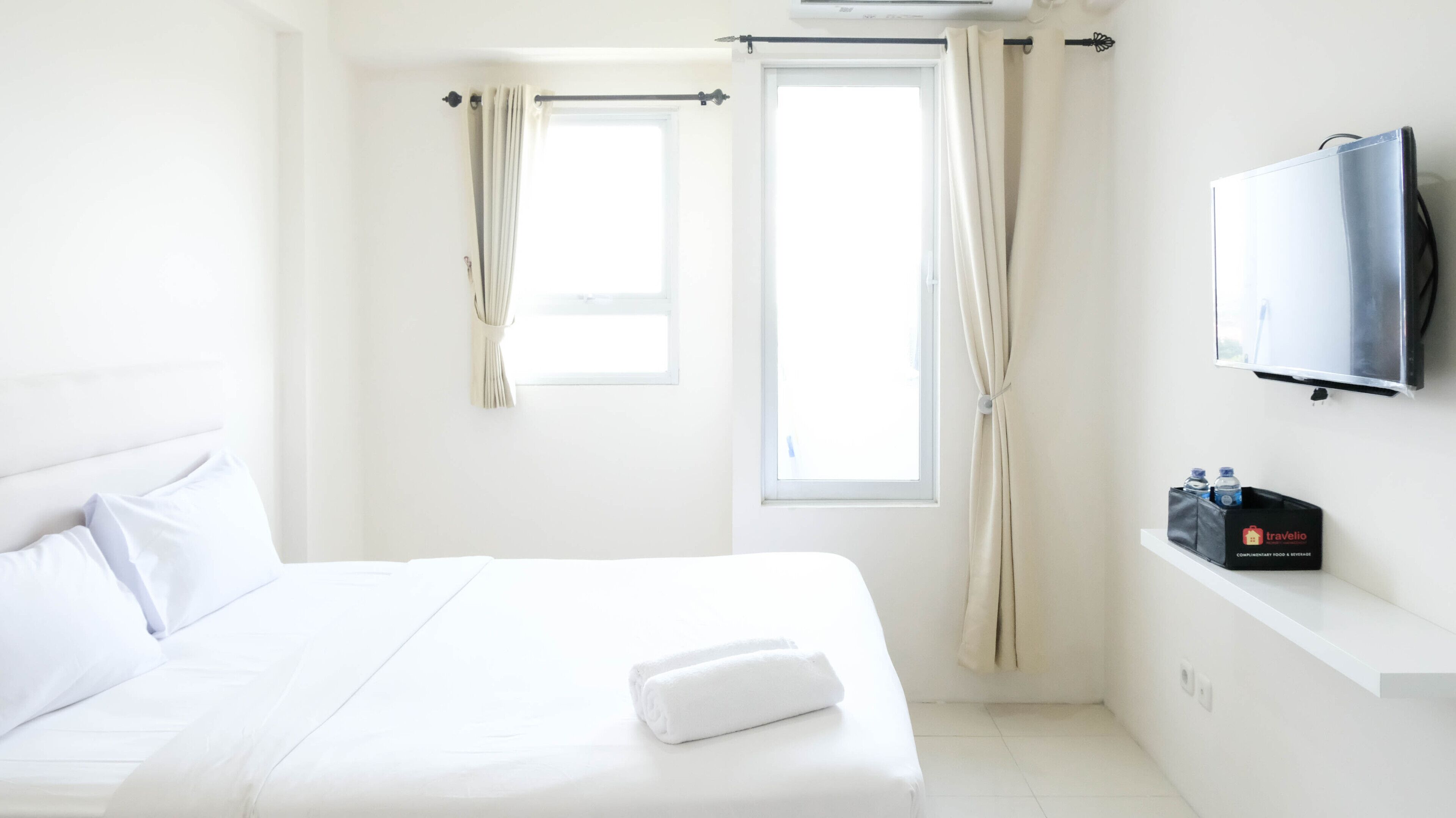 A hotel room with a bed, a TV mounted on the wall, a window with curtains, and a shelf with a travel bag and water bottles.