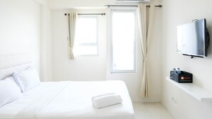 A hotel room with a bed, a TV mounted on the wall, a window with curtains, and a shelf with a travel bag and water bottles. - Best Choice And Cozy Stay Studio At Puncak Kertajaya Apartment (Surabaya)