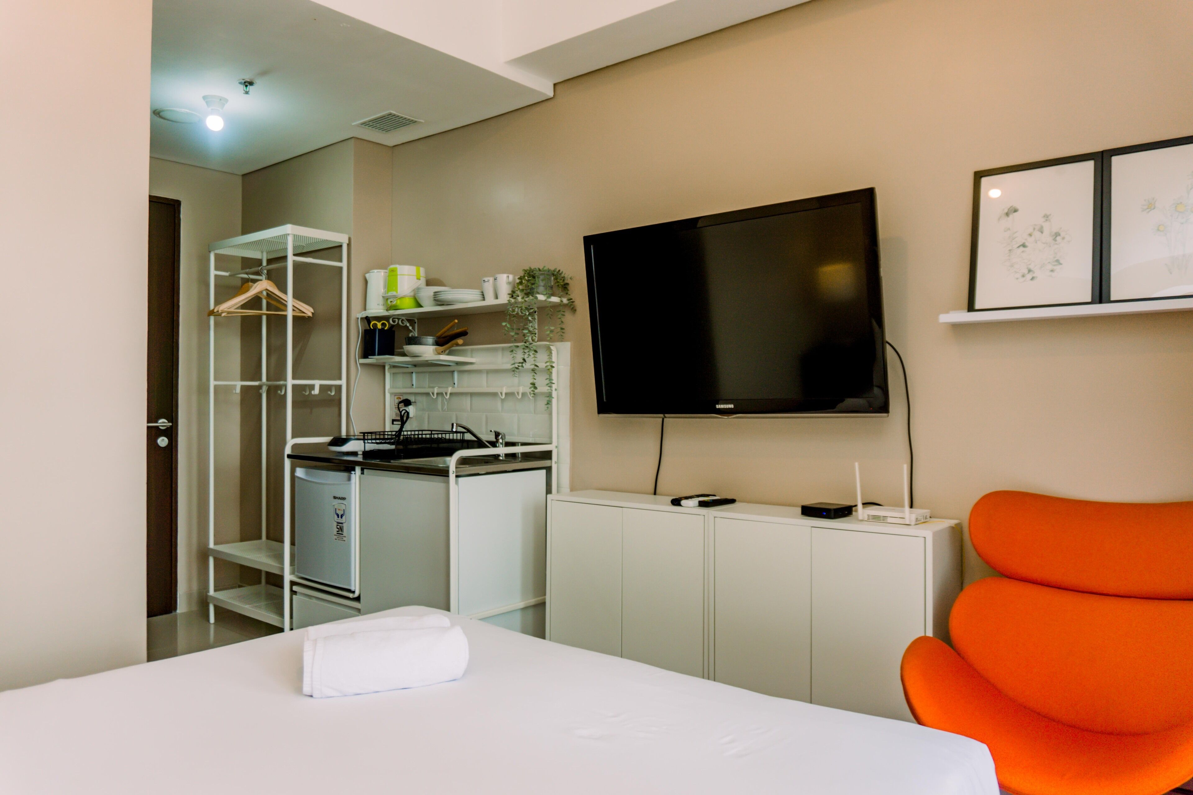 A hotel room with a bed, a TV, a kitchenette, and a bright orange chair.