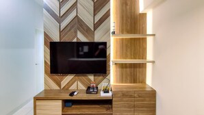 A modern living room with a large flat-screen TV mounted on a wooden wall with a chevron pattern. There are wooden shelves below the TV and a small cabinet with a remote control and a bottle on it.
