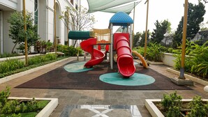 A playground with slides and a climbing structure, surrounded by a landscaped garden and a building with arched windows. - Comfy 1Br At Bellezza Apartment (Jakarta)