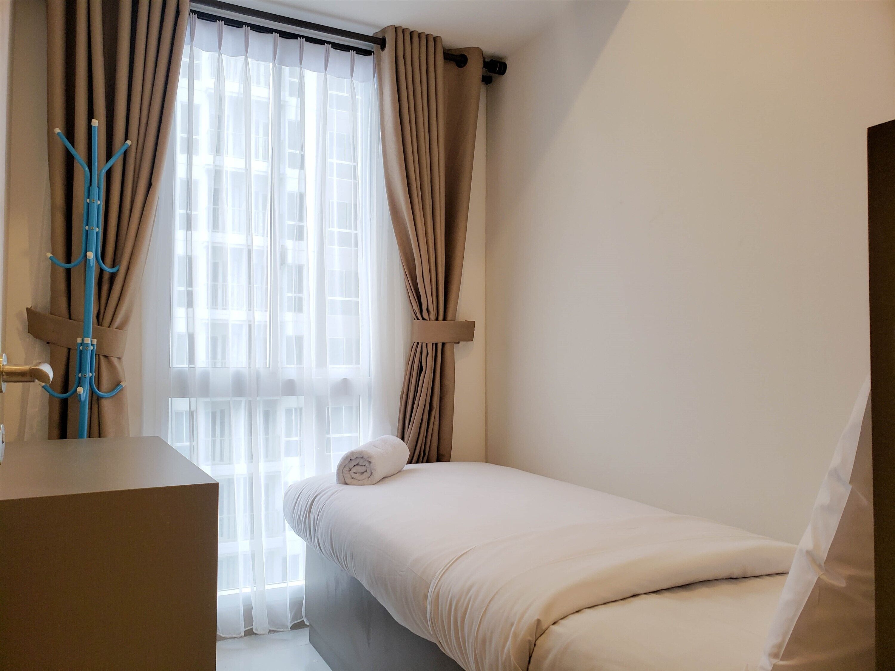 Een kleine, verzorgde slaapkamer met een bed, een dressoir en een raam met gordijnen.