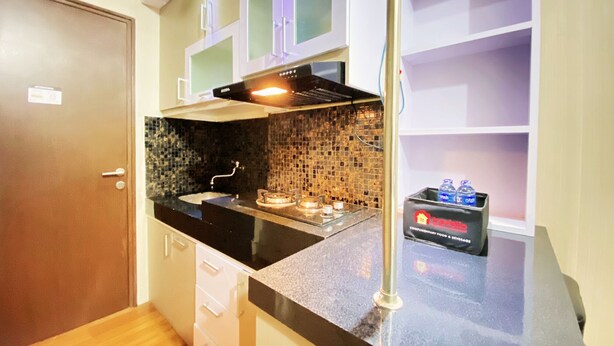 A compact kitchen with a black countertop, a stainless steel sink, and a tiled backsplash. There are white cabinets with glass doors, a stainless steel range hood, and a small box of toiletries on the countertop. - Relaxing Studio Room At Suites @Metro Apartment (Bandung)
