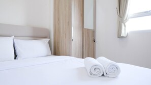 A neatly made bed with white linens and two rolled towels, a wooden headboard, and a window with curtains. - Modern And Homey 2Br At Puncak Kertajaya Apartment (Surabaya)