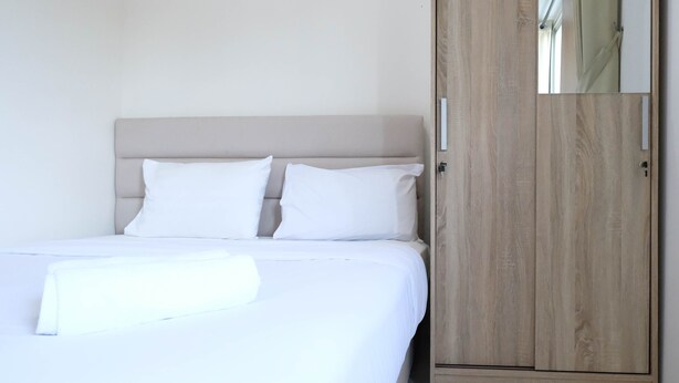 A neatly made bed with white linens and pillows, a wooden wardrobe with a mirror, and a plain wall. - Modern And Homey 2Br At Puncak Kertajaya Apartment (Surabaya)