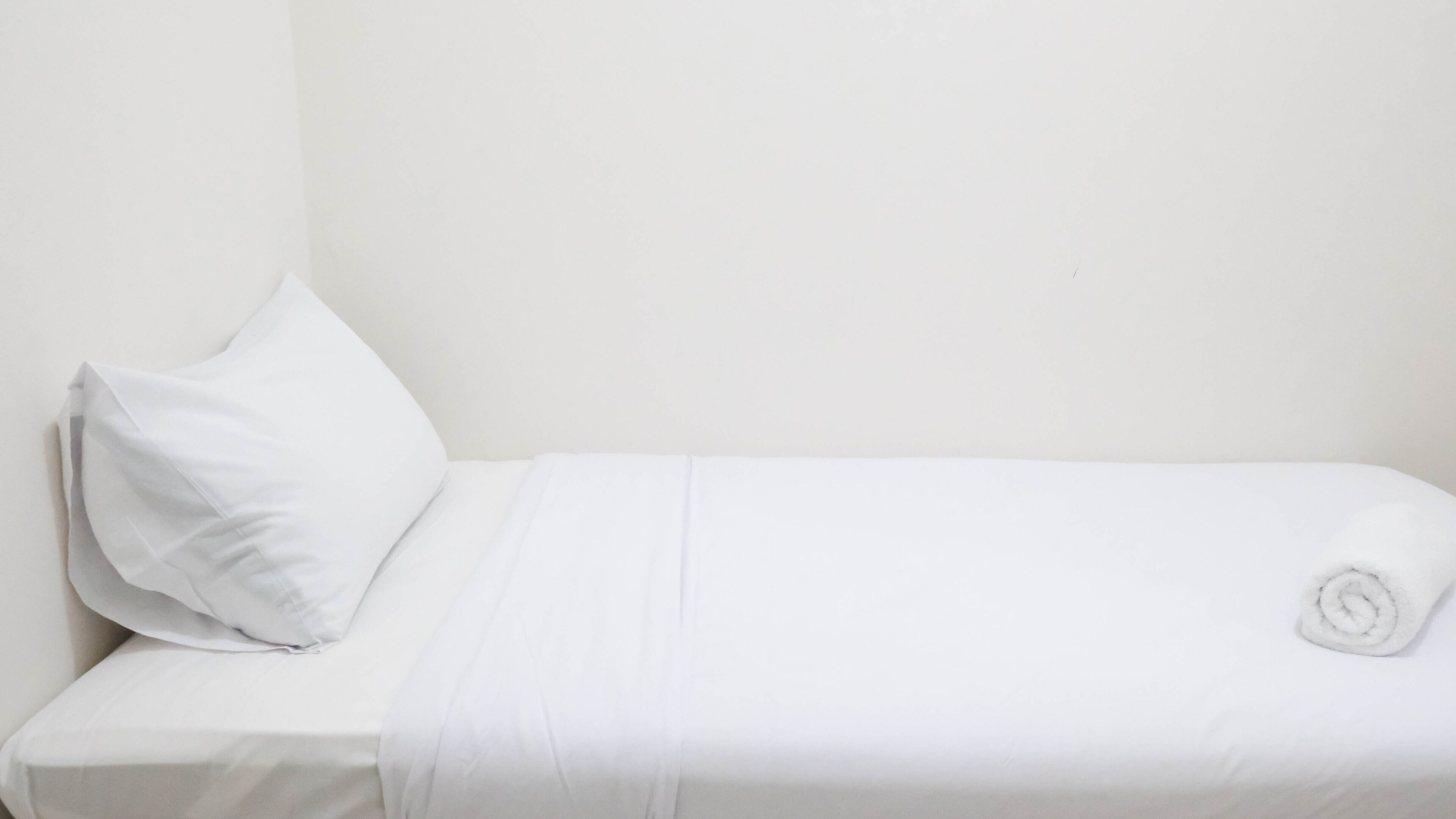 A single white bed with a pillow and a rolled towel.