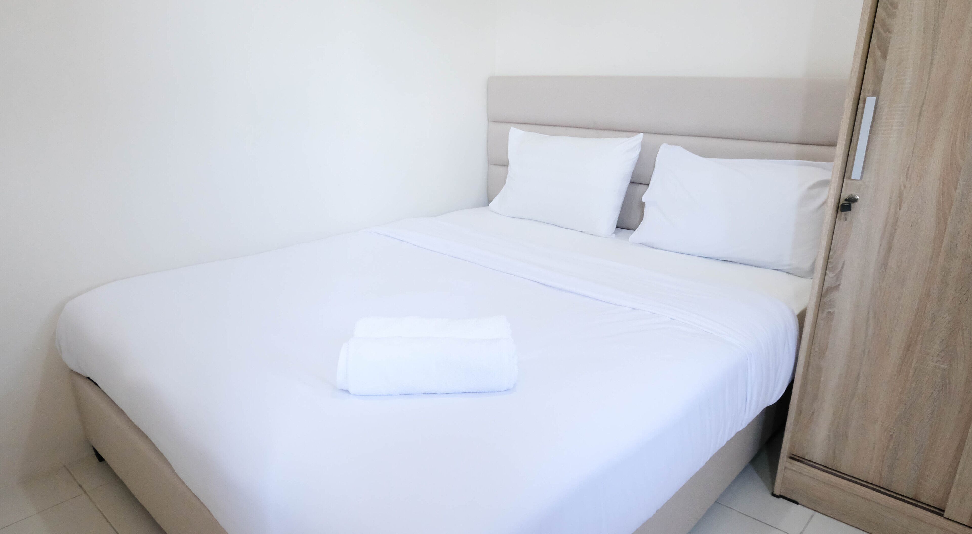Een net opgemaakt bed met witte lakens en kussens, een houten garderobe aan de rechterkant, en een schoon, minimalistisch interieur.