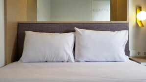 Une chambre d’hôtel comprenant un lit, deux oreillers, une tête de lit, un appareil de climatisation, une lampe fixée au mur et une prise de courant.
