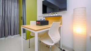 Une chambre d’hôtel avec un bureau en bois, une chaise blanche, un téléviseur à écran plat fixé au mur, une lampe et une armoire verte.