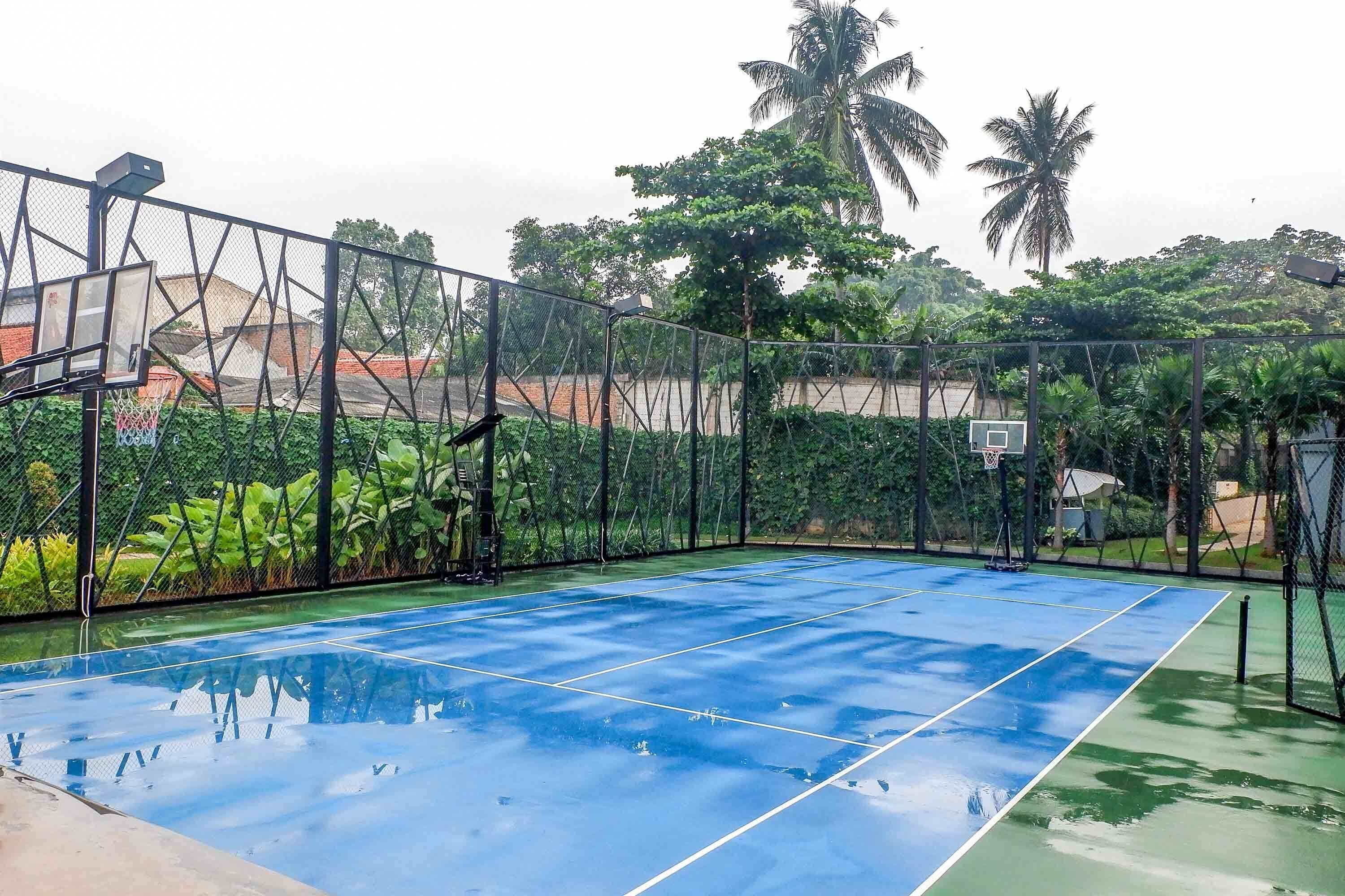 An outdoor basketball court with a blue surface, surrounded by a metal fence and greenery.