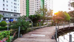 Un sentier en bois mène à un plan d’eau, qui est entour é de verdure et bordé d’un grand immeuble doté de nombreuses fenêtres.