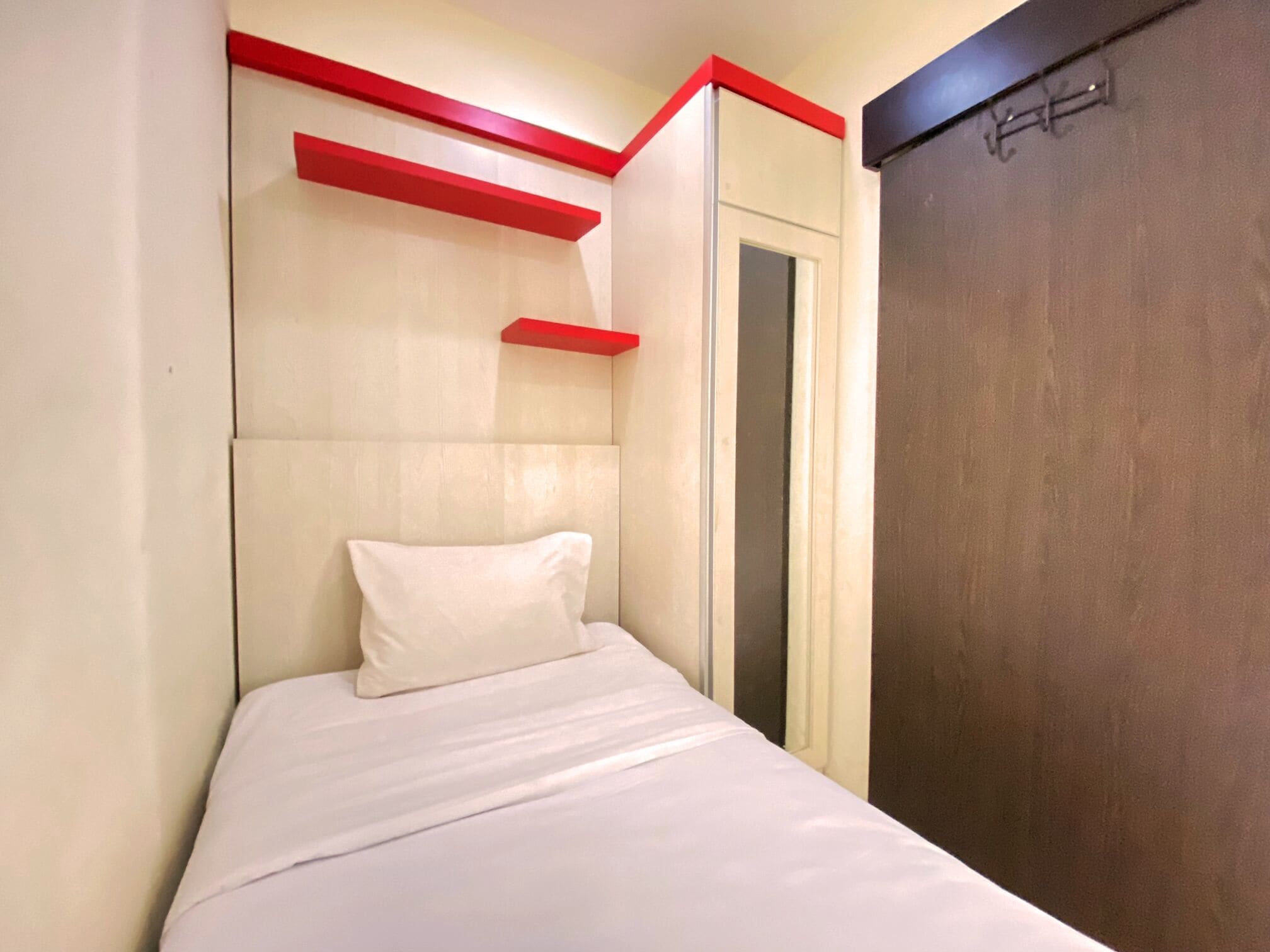 Een eenpersoonsbed met een wit matras en kussens, een houten garderobe en rode details op het hoofdeinde en de muur.