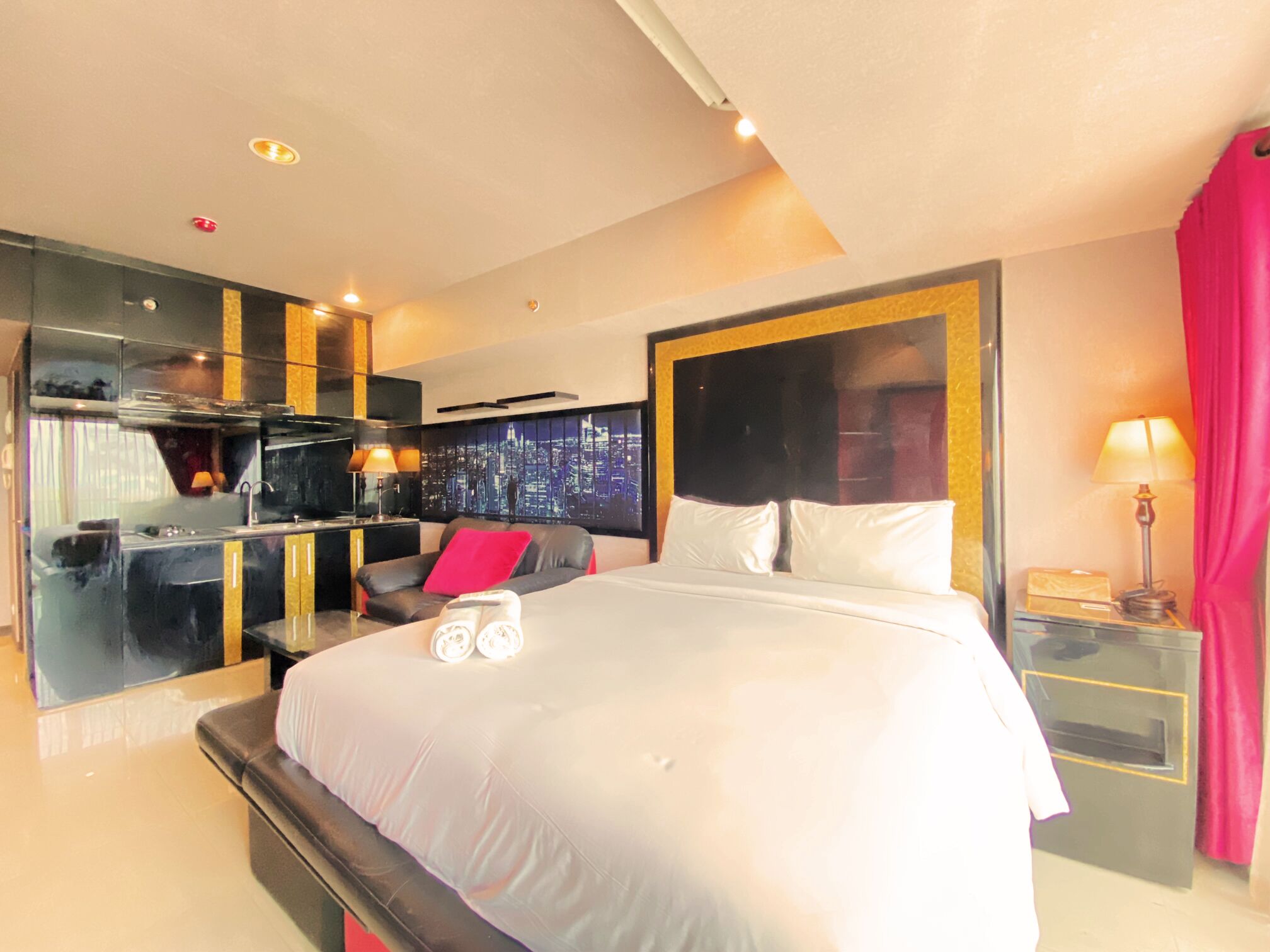 A modern hotel room with a large bed, a cityscape view, a kitchenette, and a red curtain.