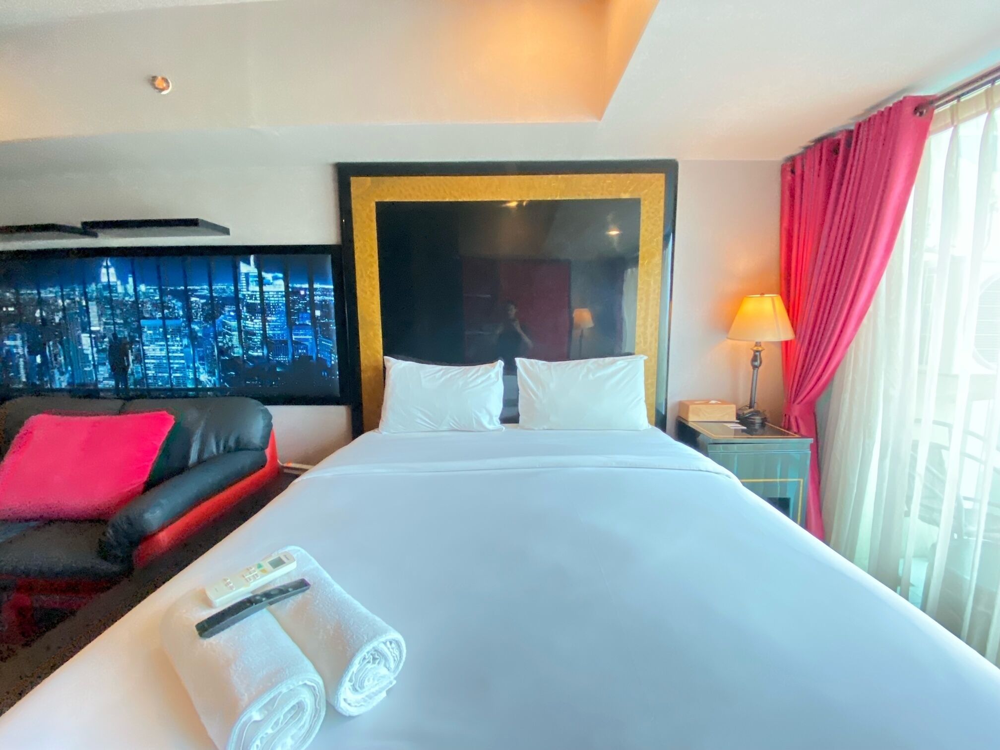 A hotel room with a large bed, a TV displaying a cityscape, a red sofa, and a lamp.