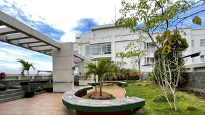A modern building with a covered outdoor area featuring a curved planter with greenery, a tiled walkway, and a view of the building's exterior. - Cozy Living 1Br Apartment At Parahyangan Residence (Bandung)