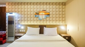 Ein Hotelzimmer mit einer gepolsterten Kopfteile, einem Bett mit weißen Bettwäsche, zwei Nachttischen und einem Wandbild, das einen Sonnenuntergang über Wasser zeigt.