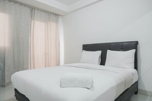 Una cama bien hecha con sábanas blancas y una toalla doblada, cabecera oscura y cortinas traslúcidas en una ventana.